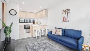 Une cuisine moderne avec des meubles blancs, un îlot central et des tabourets de bar. On y trouve un canapé bleu avec un coussin décoratif et un tapis gris sur le sol.