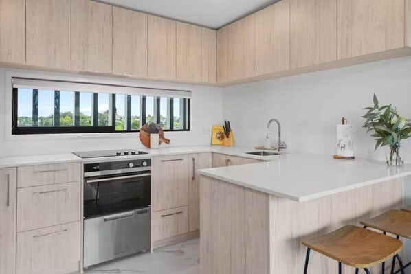 A modern kitchen with light wood cabinets, a large window, a built-in oven, a sink, and bar stools.