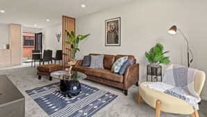 A modern living room with a brown leather sofa, a round coffee table, and a cream-colored armchair. There is a large window with a view of a brick building outside.