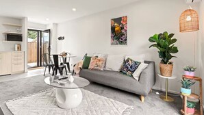 A modern living room with a grey sofa, a glass coffee table, and a dining area with a white table and black chairs. - Vibrant 2 bed Henderson Abode (Auckland)