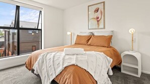 A modern bedroom with a large window, a bed with orange bedding, a white throw blanket, and two bedside tables with lamps. - Vibrant 2 bed Henderson Abode (Auckland)