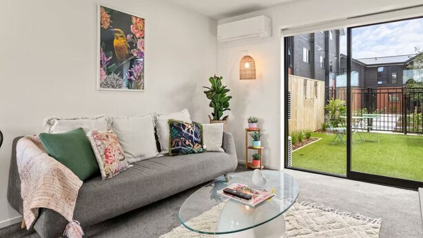 A modern living room with a grey sofa, colorful cushions, a glass coffee table, and a large window with a view of a backyard. - Vibrant 2 bed Henderson Abode (Auckland)
