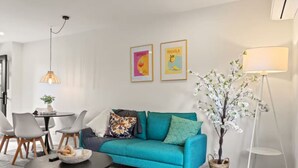 En moderne stue med en turkis sofa, et rundt sofabord og et spiseområde med stole og et lille bord. Der er en gulvlampe og en potteplante.