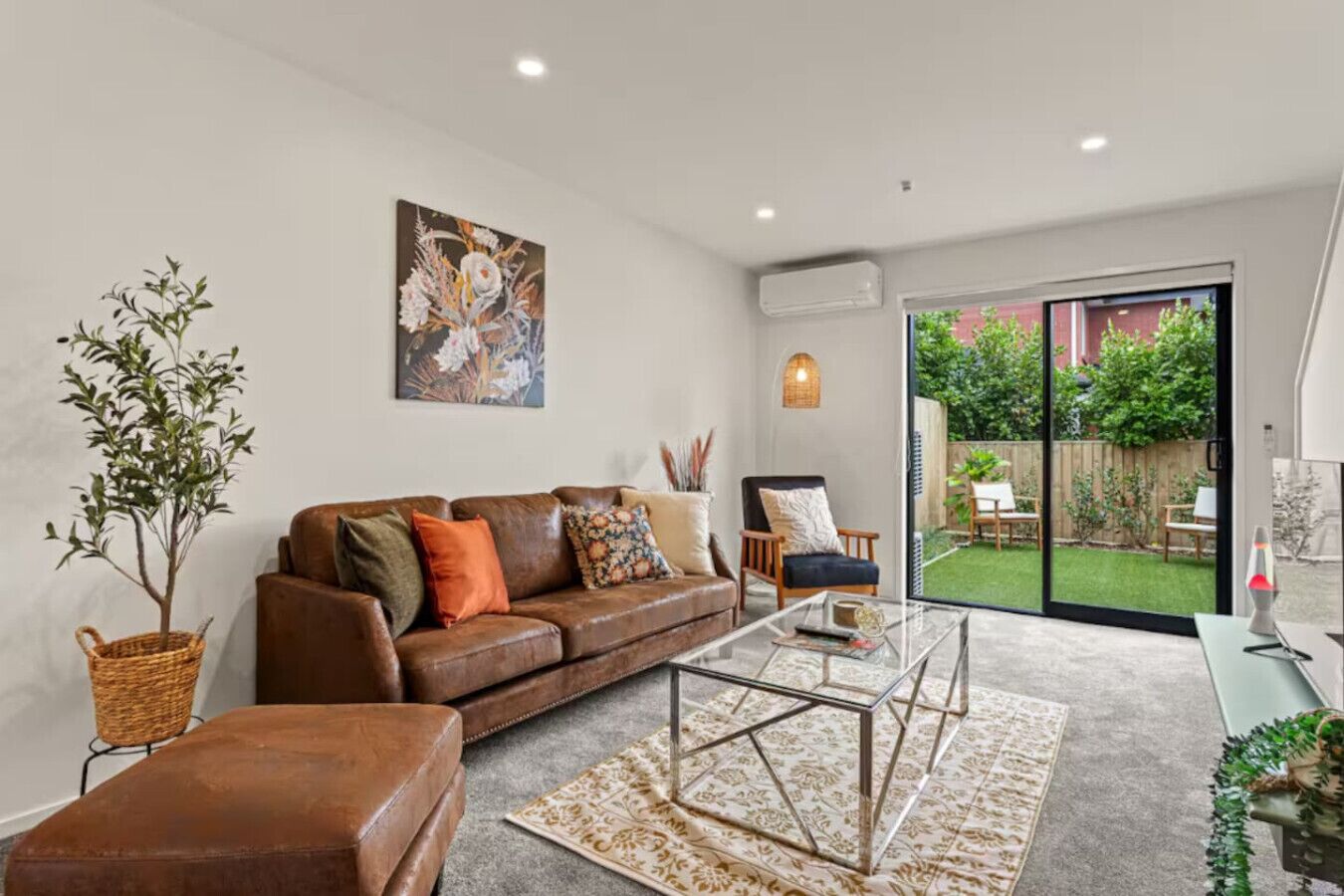 A modern living room with a brown leather sofa, a glass coffee table, and a large window offering a view of a garden.