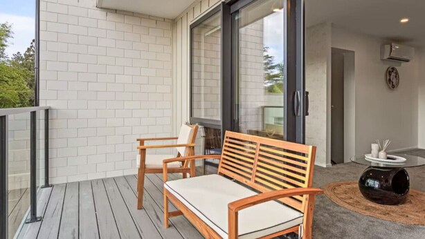 A balcony with a wooden bench and chairs, a glass door leading inside, and a small table with a decorative item. - Well-styled Vibrant 2 Bed Home (Auckland)