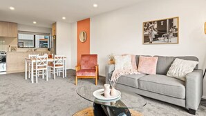A modern living room with a grey sofa, a red armchair, and a glass coffee table. There is a dining area with a white table and chairs, and a kitchen in the background. - Well-styled Vibrant 2 Bed Home (Auckland)