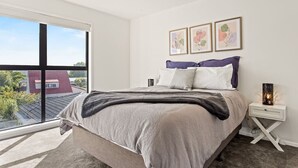 A modern bedroom with a large window, a bed with a grey and white duvet, a nightstand with a lamp, and three framed floral artworks on the wall. - Central Auckland Townhouse (Auckland)