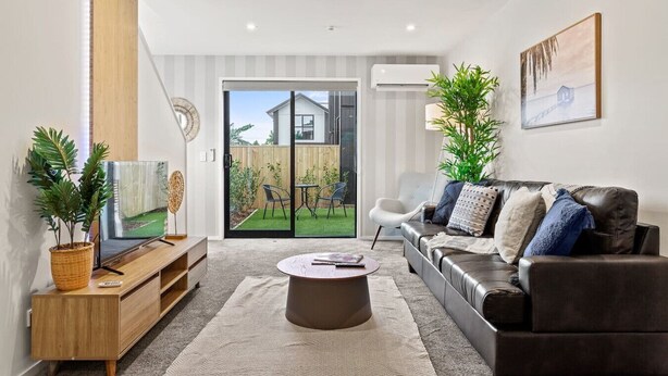 Ein modernes Wohnzimmer mit Ledersofa, Couchtisch und Fernseher auf einem Holzboden. Eine Glastür öffnet sich zu einem Garten mit Terrasse, im Hintergrund ist ein Haus zu sehen.