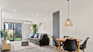 Ein modernes Wohnzimmer mit einer Couch, einem Couchtisch und einem Esstisch mit Stühlen. Eine Glastür führt zu einem Balkon mit Pflanzen.