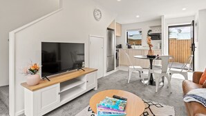 Un salon moderne avec un téléviseur à écran plat posé sur un meuble en bois, un coin repas avec une table et des chaises, et une cuisine en arrière-plan.