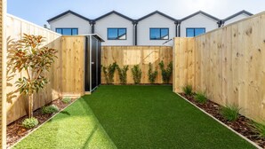A modern residential building with a fenced backyard, artificial grass, and a row of small trees.