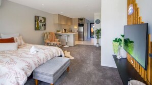 A modern bedroom with a large bed, a small seating area, a kitchenette, and a TV mounted on the wall. - Cozy Studio on Macky Street (Lower Hutt)