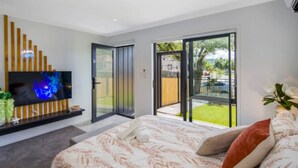 A modern bedroom with a large bed, a TV mounted on a wooden panel, and a sliding glass door leading to a balcony. - Cozy Studio on Macky Street (Lower Hutt)