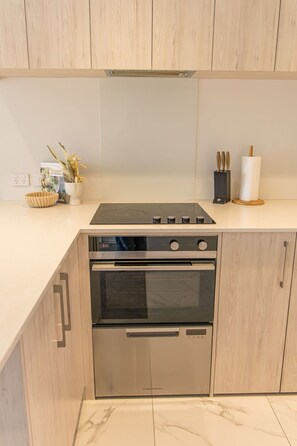 A kitchen with a built-in stove, oven, and microwave. There is a white countertop, wooden cabinets, and a small plant on the counter. - Cozy Studio on Macky Street (Lower Hutt)