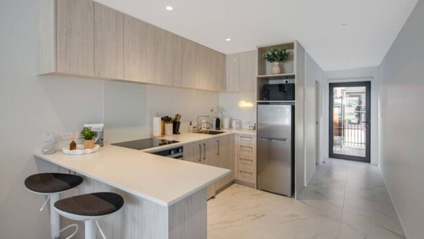 A modern kitchen with a central island, bar stools, a built-in oven, and a stainless steel refrigerator. - Cozy Studio on Macky Street (Lower Hutt)