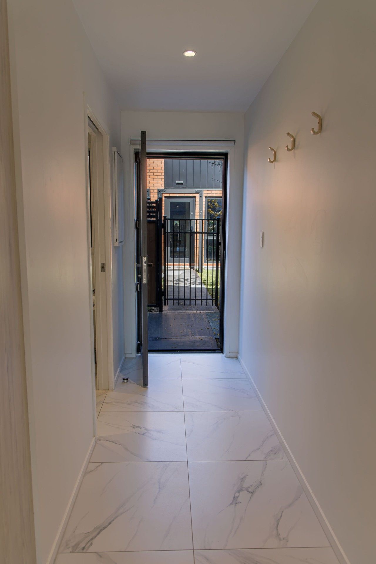 Un couloir avec une porte donnant sur une entrée sécurisée par un portail, un sol carrelé et un luminaire fixé au mur.