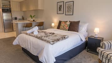 A modern bedroom with a large bed, a nightstand, a chair, and a kitchenette in the background.