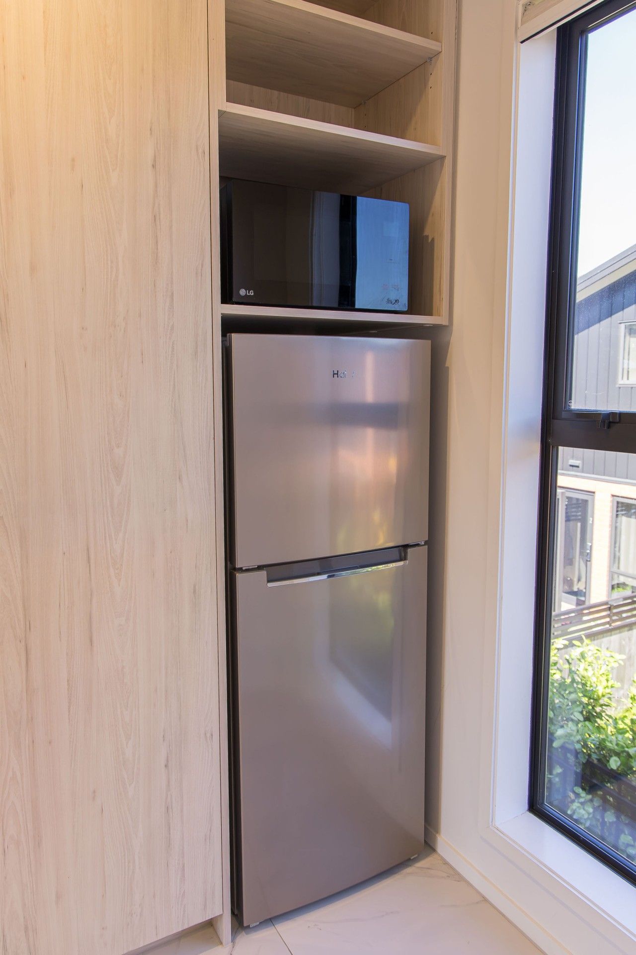 A compact kitchen with a built-in stainless steel refrigerator, a window with a view of the outside, and a wooden cabinet above the fridge.