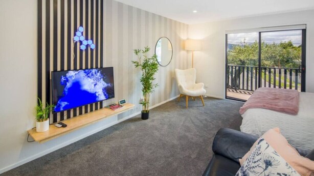 A modern hotel room with a large TV, a comfortable chair, a bed, a balcony with a view, and a potted plant. - Bright Studio Apartment on Macky Street (Lower Hutt)