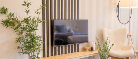 A modern living room with a flat-screen TV mounted on a wall, a white armchair, a round coffee table, and a potted plant.