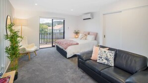 A modern bedroom with a large bed, a black leather sofa, a small white chair, and a balcony with a view. - Bright Studio Apartment on Macky Street (Lower Hutt)
