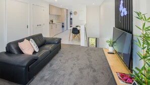A modern living room with a black leather sofa, a flat-screen TV, and a grey carpet. - Bright Studio Apartment on Macky Street (Lower Hutt)