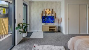 A modern bedroom with a large TV, a wooden entertainment unit, and a sliding glass door leading to a balcony. - Macky Street Studio Retreat (Lower Hutt)