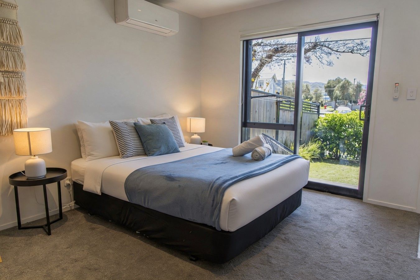 A bedroom with a bed, two bedside tables with lamps, a large window with a view of the outdoors, and a ceiling air conditioning unit.
