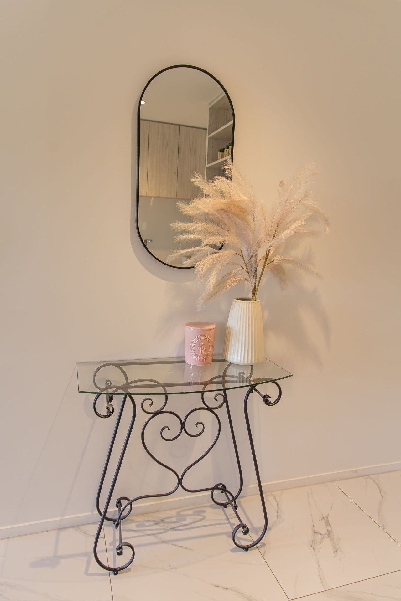 A glass console table with a mirror above it, a vase with dried pampas grass, and a pink candle.