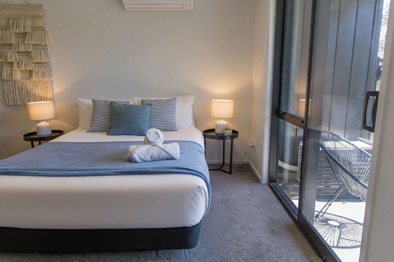 A hotel room with a large bed, two bedside lamps, a wall-mounted macrame piece, and a view of the outside through a glass door.