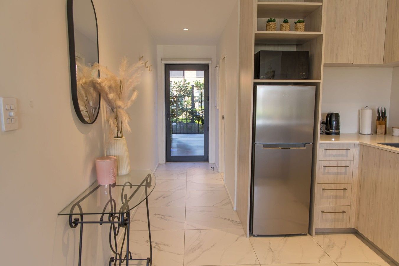 A modern kitchen with a stainless steel refrigerator, wooden cabinets, and a view through the door to an outdoor area.
