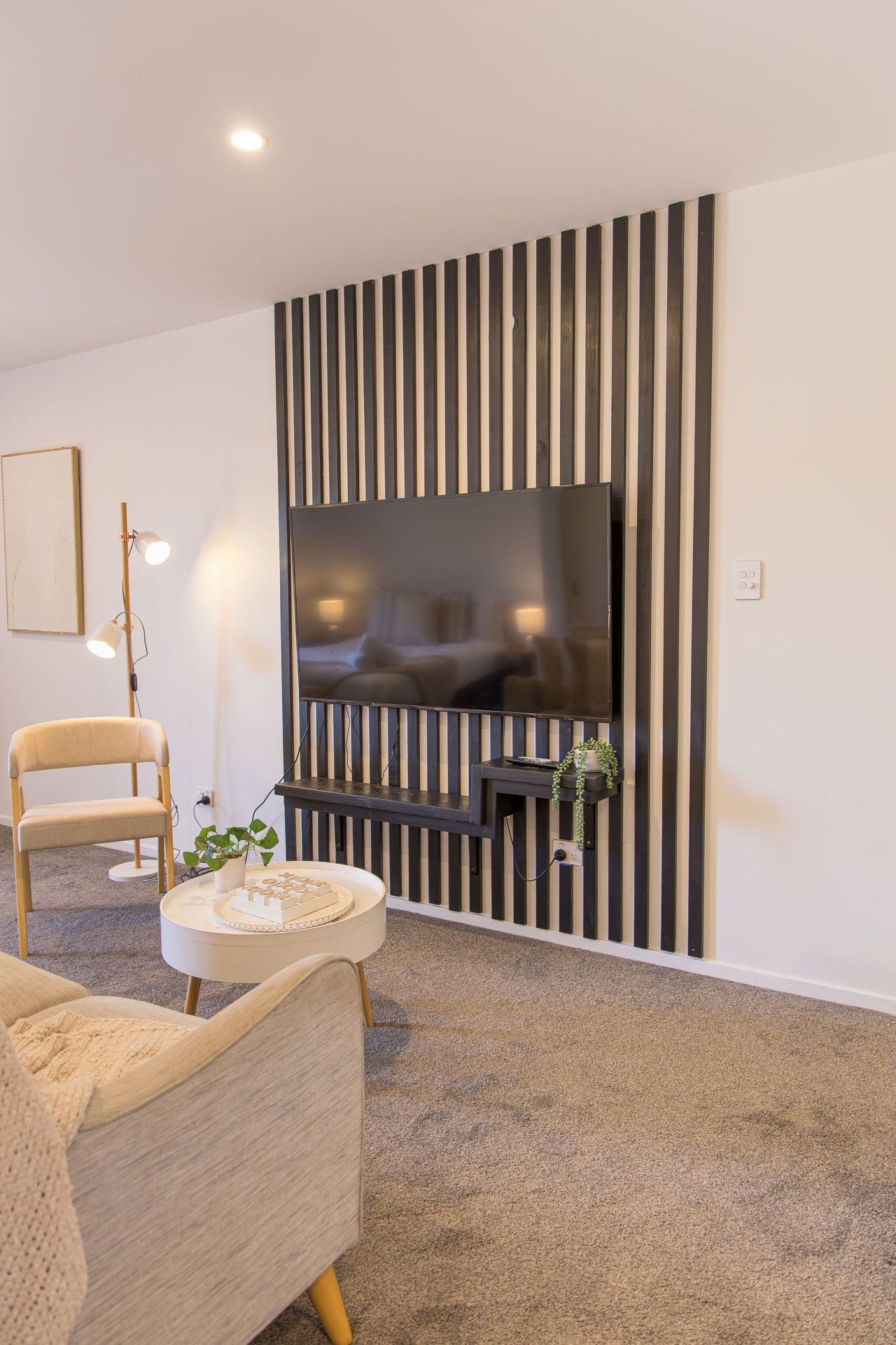 A modern living room with a flat-screen TV mounted on a black and white striped wall, a round coffee table with a plant, and a beige armchair.