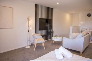 A modern living room with a sofa, a small round table, a floor lamp, and a flat-screen TV mounted on a wall with vertical blinds. - Peaceful Hideaway: Studio in Macky (Lower Hutt)