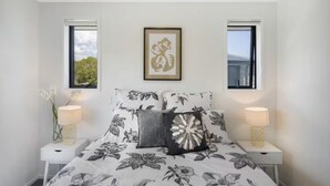 A bedroom with a floral bedspread, two bedside lamps, a framed artwork above the bed, and windows with views of greenery. - Your Slice of Heaven in the Hutt (Lower Hutt)