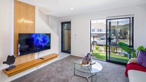 A modern living room with a flat-screen TV mounted on a wooden panel, a glass coffee table, and a sofa with a red throw. - Your Slice of Heaven in the Hutt (Lower Hutt)