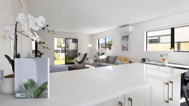 A modern kitchen with white cabinetry, a central island, and a dining area with a gray sofa and a small table. - Homely Haven in Trentham - 2BR (Upper Hutt)