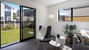 A modern living room with a sliding glass door leading to a garden, a black armchair, a round white side table, and a grey sofa with a floral cushion. - Homely Haven in Trentham - 2BR (Upper Hutt)