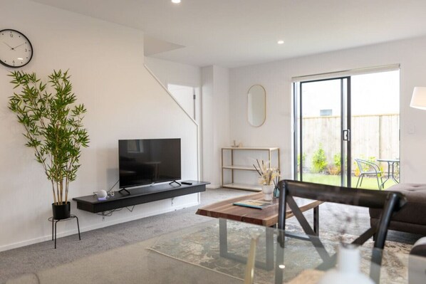 Una sala de estar moderna con un televisor de pantalla plana sobre un mueble negro, una planta grande en maceta, una mesa de centro de madera y un enorme ventanal corredizo que da a un patio.