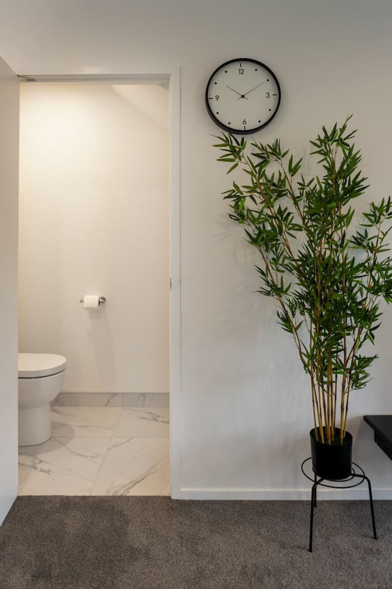 A bathroom with a toilet, a clock on the wall, and a bamboo plant on a stand.