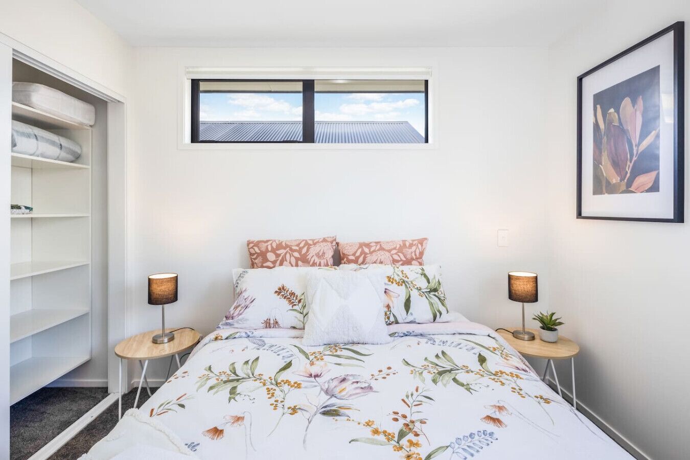 Une chambre à coucher comprenant un lit, deux tables de chevet, une fenêtre, un tableau encadré au mur et une armoire avec des étagères.