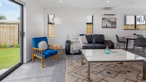 A modern living room with a glass coffee table, a blue armchair, a black leather sofa, and a dining area with a small table and chairs. - Scenic Views from our 2-BR Trentham Hideaway (Upper Hutt)