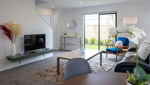 A modern living room with a flat-screen TV, a coffee table, a sofa, and a chair. There is a staircase leading to another level, and a large window providing a view of the outdoors. - Scenic Views from our 2-BR Trentham Hideaway (Upper Hutt)