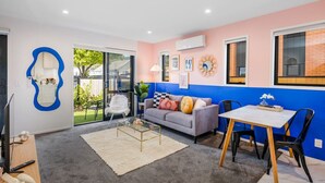 A modern living room with a grey sofa, a coffee table, and a dining table with chairs. There is a large mirror on the left wall and a blue accent wall. - Hutt Hideaway on Milne (Lower Hutt)