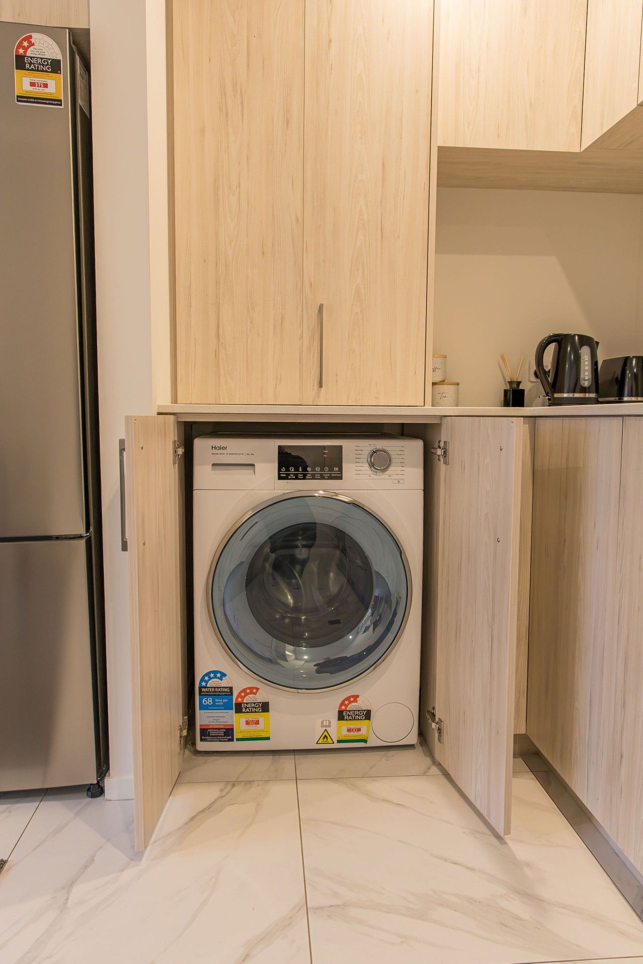 Una unidad compacta de lavadora y secadora con calificaciones de eficiencia energética y de agua, ubicada en una cocina con armarios de madera y piso de baldosas.