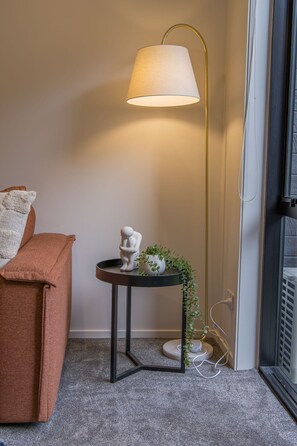 A room with a brown sofa, a small round table with a lamp, and a white sculpture. - Groveview Oasis: 1BR Retreat (Lower Hutt)