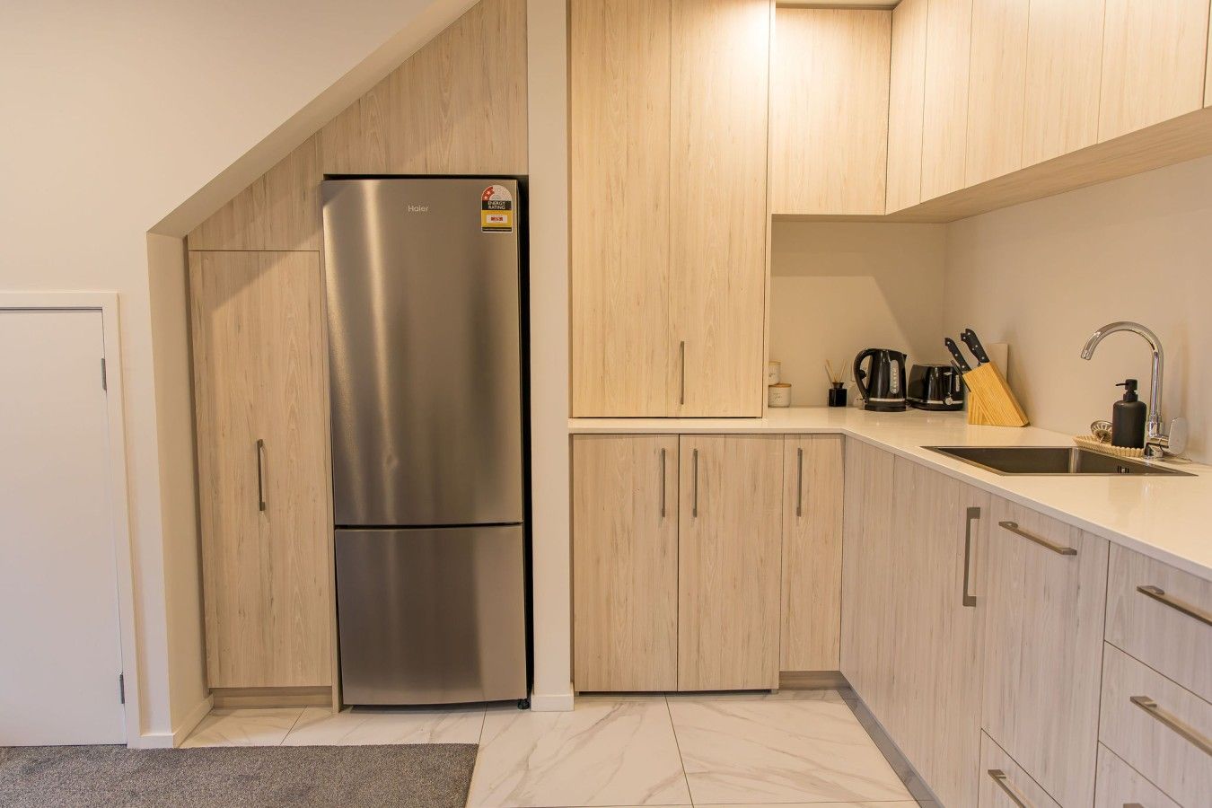 A modern kitchen with a stainless steel refrigerator, wooden cabinets, a countertop sink, and various kitchen utensils.