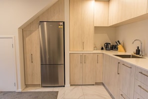 A modern kitchen with a stainless steel refrigerator, wooden cabinets, a countertop sink, and various kitchen utensils. - Groveview Oasis: 1BR Retreat (Lower Hutt)