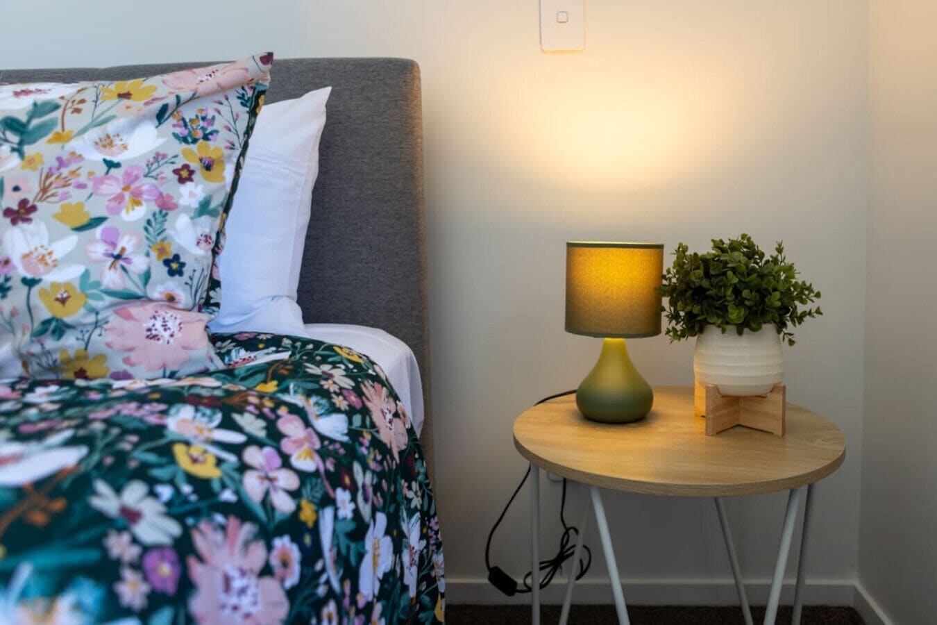Ein Bett mit floraler Bettwäsche, eine Nachttischlampe und ein Blumentopf auf einem kleinen Holztisch neben dem Bett.