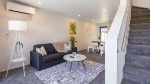 A modern living room with a sofa, coffee table, and floor lamp. There is a staircase to the right and a dining area in the background. - Ariki Shire: A Wellington Fantasy (Lower Hutt)