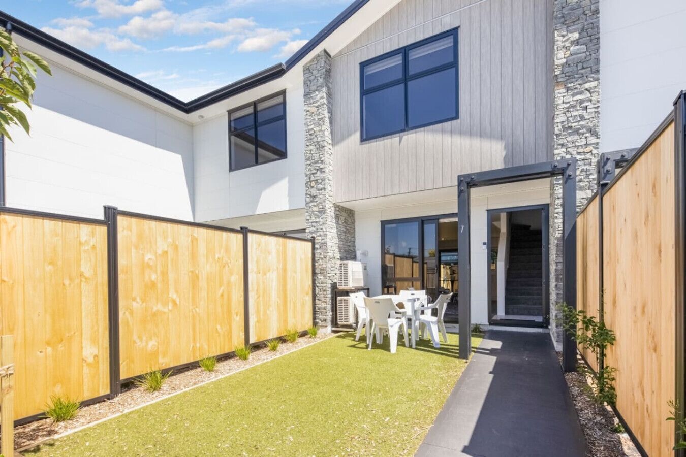 Une maison moderne à deux étages, dotée d’une façade en pierre, d’une cour arrière clôturée et d’un patio aménagé avec du mobilier d’extérieur.